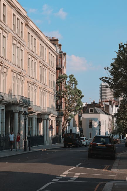 A street scene in a residential area showing a line of classical-style Victorian buildings with beige facades and black wrought iron balconies on the left side of the image, set against a bright blue sky with some scattered clouds. In the foreground, a black van is parked on the pavement while another vehicle is slightly further along the street. Several cardboard boxes, wrapped furniture pieces in plastic, and blankets are arranged on the sidewalk near the building entrance, suggesting preparations for a home relocation. A hand truck, or dolly, is visible near the boxes, and a person is seen carrying a box towards a nearby vehicle suggesting active loading or unloading. The environment is well-lit, indicating daytime, and there are some trees with green foliage along the street, providing shade. The scene aligns with professional removals services such as those offered by Man and Van Bayswater, specialising in furniture transport and packing and moving logistics for local house relocations near Queensway.