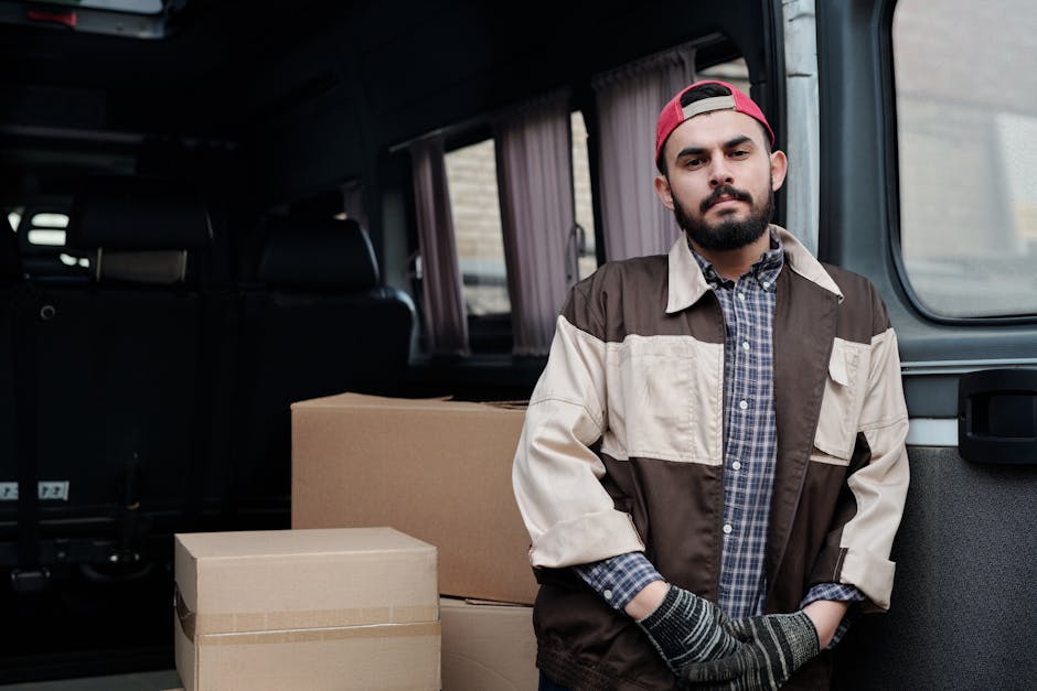A man with a beard and wearing a red cap, brown and beige jacket, and gloves stands inside a van during a house removal process. Behind him, there are two cardboard moving boxes, one large and one smaller, placed on the vehicle's floor. The van's interior has grey walls and seats, with a window showing a blurred outdoor scene. The Man and Van Bayswater team appears to be preparing for furniture transport and home relocation, with the man positioned near the open rear of the vehicle, ready to load or unload belongings as part of the packing and moving service. The scene captures the loading process, including the materials used for packing, such as cardboard boxes, within a professional setting focused on efficient and careful house removals, supporting the website's focus on same-day moving near Queensway.