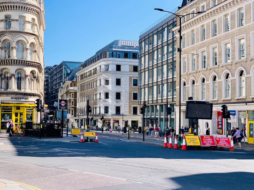The image shows a city street scene with a large, multi-story residential or commercial building constructed from brown brick with white window frames and balconies along the upper floors. In the fore