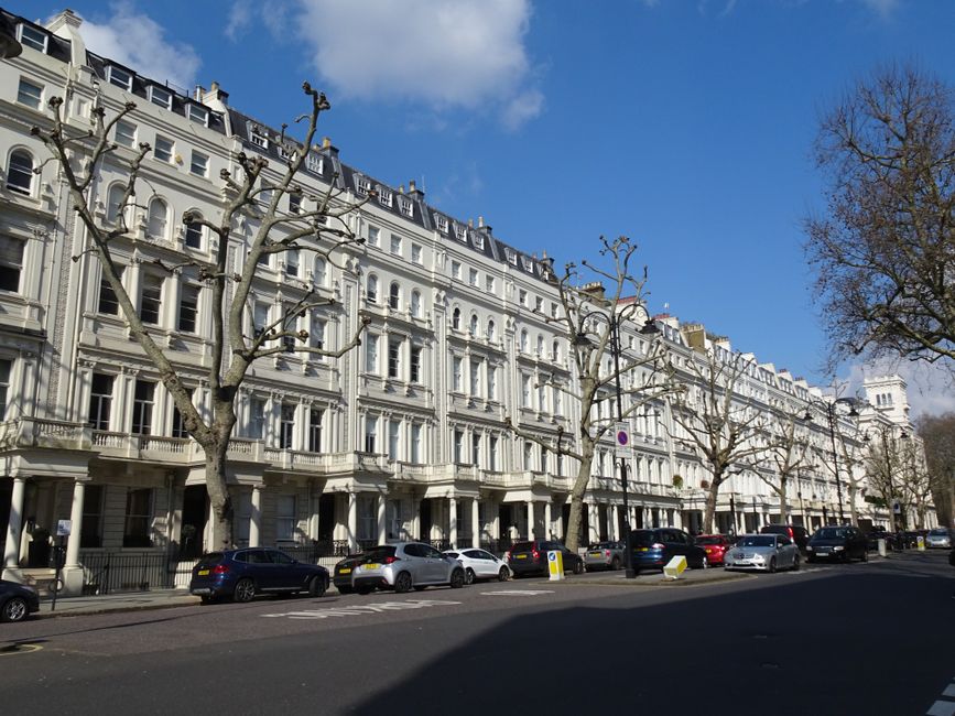 A daytime exterior view of a row of elegant white Victorian-style terraced buildings lining a street in Bayswater, Westminster, with leafless trees standing in front and a clear blue sky overhead. Sev