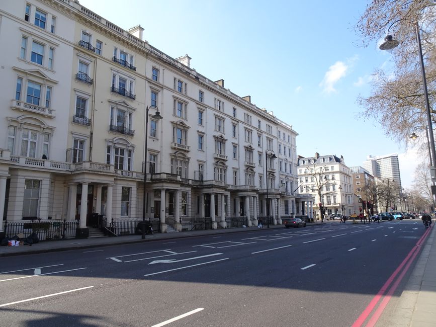 A daytime exterior view of a row of elegant white Victorian-style terraced buildings lining a street in Bayswater, Westminster, with leafless trees standing in front and a clear blue sky overhead. Sev